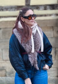 Veste en cuir noire avec un col en fausse fourrure, pull beige avec cordons de serrage, et jean bleu ; des lunettes de soleil surdimensionnées complètent le look.