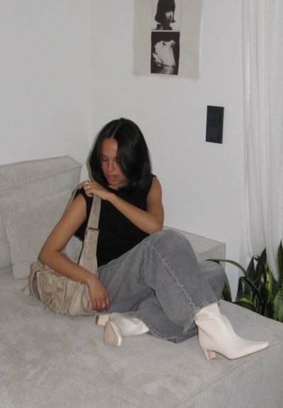 Bolso beige, top sin mangas negro, pantalones anchos grises, botas blancas hasta el tobillo con tacones cuadrados. Sentada en un sofá de textura clara.