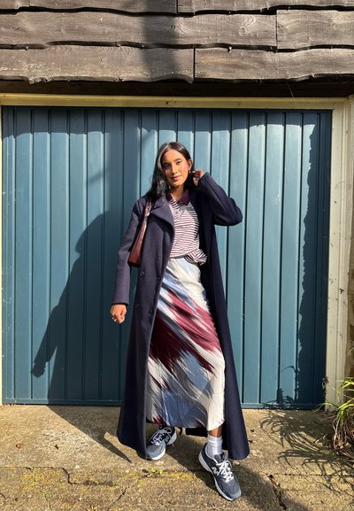 Abrigo de lana azul marino, camiseta de rayas y una falda larga con un patrón rojo y blanco. Combinado con zapatillas grises, de pie contra una puerta de madera azul.