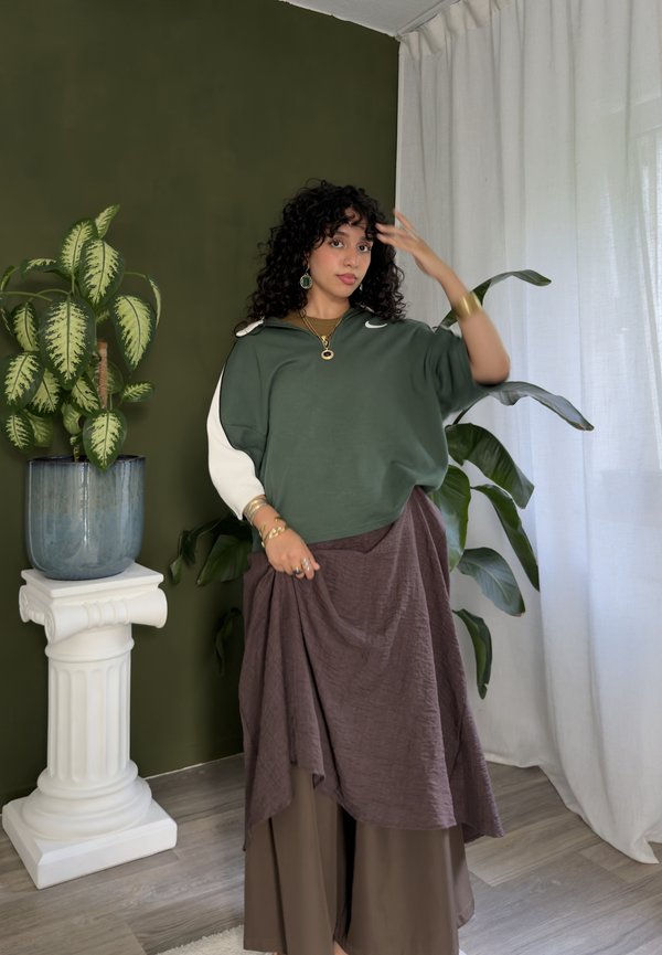 Femme aux cheveux bouclés se tient à l'intérieur près de plantes et de rideaux, portant un haut vert et blanc avec une jupe marron superposée et des bijoux en or.