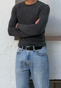 Άνδρας με σταυρωμένα χέρια, φοράει μπλούζα μακρυμάνικη σκουρόχρωμη γκρι, ανοιχτόχρωμα μπλε τζιν και μαύρη ζώνη, στέκεται μπροστά σε έναν λευκό τοίχο με υφή.