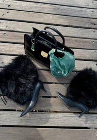 Svart skinnveske med gullhardware, teal cap, og svarte faux fur ankelstøvler med spiss hæl på en treoverflate.