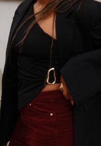 Schwarzes eng anliegendes Oberteil mit gedrehtem Detail, metallischem ovalem Akzent an der Seite, getragen unter einem schwarzen Blazer, kombiniert mit burgunderroten, hoch taillierten Hosen.