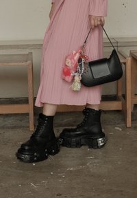 Botas de plataforma negras, gruesas, con cordones y acentos metálicos. Acompañadas de un bolso negro con llaveros coloridos y un juguete de peluche adjunto.