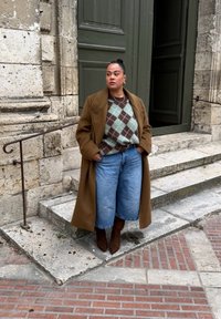 Manteau marron oversize, pull à losanges verts et marron, culottes en jean bleu clair, bottes à talons marron, posée près d'escaliers en pierre et d'une porte.