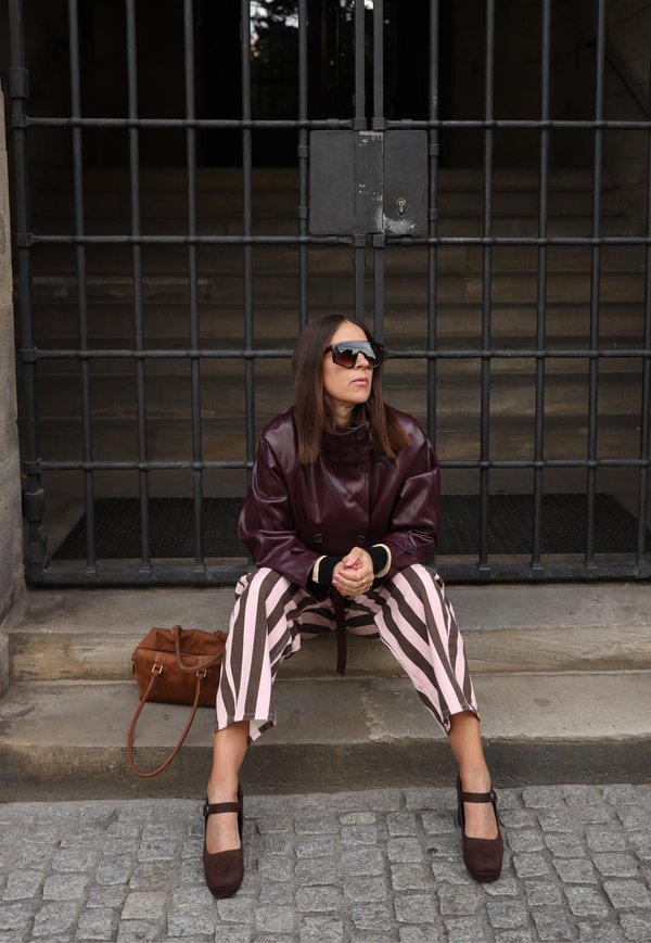 Mujer con gafas de sol, chaqueta granate y pantalones a rayas sentada en un escalón de piedra frente a una gran puerta metálica negra con un bolso marrón a su lado.