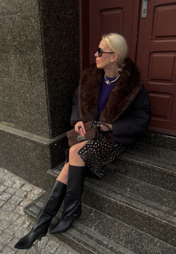 Une femme blonde portant des lunettes de soleil est assise sur des marches en pierre près d’une porte rouge. Elle porte un manteau à col de fourrure et de hautes bottes noires.