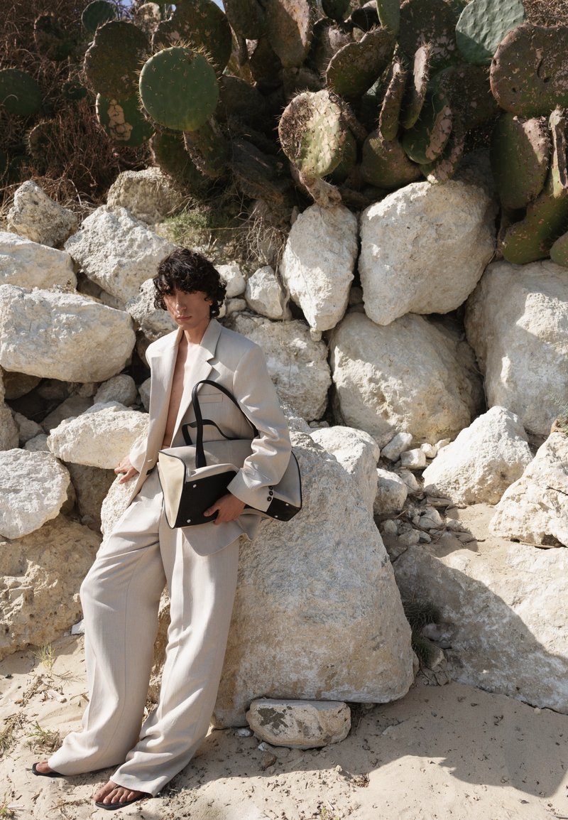 Personne en costume beige tenant un sac à main, debout pieds nus sur un sol sablonneux près de grosses pierres et de plantes de cactus.