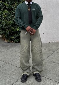 Green sweatshirt with a crew neck, black tie, light grey wide-leg trousers featuring curved seams, and black shoes with fringe detailing.