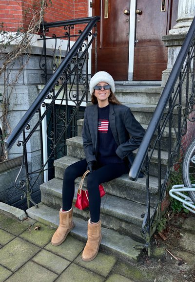 Mujer con botas marrones, leggings negros, blazer gris, sombrero de piel blanca y gafas de sol, sentada en unos escalones de piedra al aire libre sosteniendo un bolso rojo.