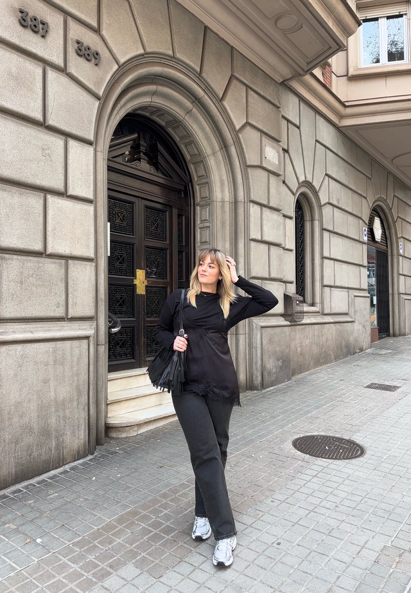 Donna in abito nero e scarpe da ginnastica bianche in piedi sul marciapiede di una città vicino a un edificio in pietra con porte e finestre ad arco in legno.