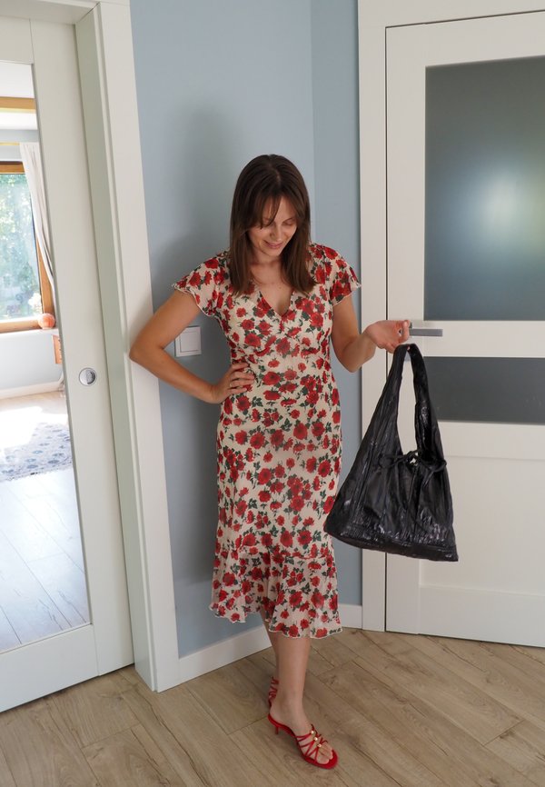 Femme en robe fleurie et talons rouges tenant un sac à main noir, debout à l'intérieur près d'une porte blanche et d'un mur peint en bleu clair.