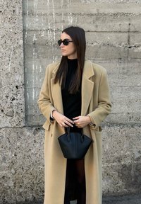 Beež villane mantel, must topp ja mini-must käekott. Pikk tumedad juuksed, ümmargused mustad päikeseprillid, seisab tekstuurse betoonseina vastu.