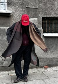 Dolga črna bunda z rjavim šalom, burgundska bluza, črne hlače in rjave čevlje. Rdeča kapa z belim napisom. Siva teksturirana stena v ozadju.