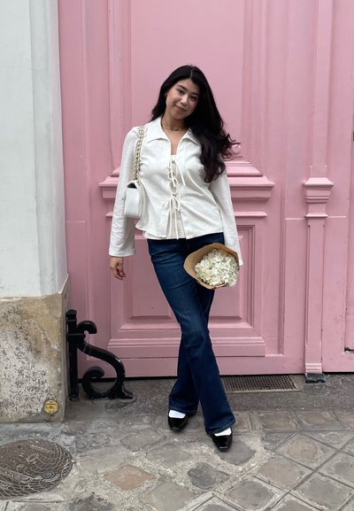 Blusa blanca con cordones, jeans de campana azules, zapatos negros con acentos blancos. Sosteniendo un ramo de flores blancas frente a una pared rosa.
