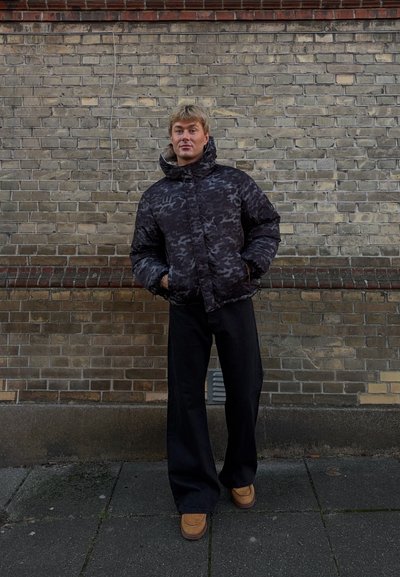 Chaqueta acolchada de camuflaje negra con cuello alto, combinada con pantalones negros de pierna ancha y zapatos mocasines color canela, con una pared de ladrillos como fondo.