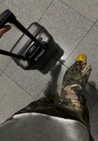Geantă de voiaj neagră, dură, cu mânere duale extensibile, plasată pe un podea gri cu plăci. Pantaloni de camuflaj și pantofi galbeni, parțial vizibili.