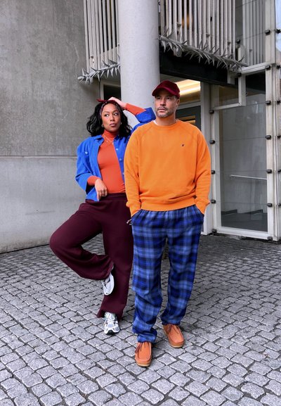 Dos personas posan al aire libre. Una lleva una chaqueta azul, una camiseta naranja, pantalones burdeos y zapatillas blancas. La otra lleva un sudadera naranja y pantalones de cuadros.