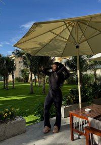 Kvinne i svart antrekk og beige klippere justerer hodetelefoner under en stor beige parasoll nær et bord og palmer på en solfylt dag.