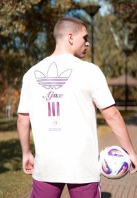 Tricou alb cu un logo Adidas violet și textul "Ajax", având trei dungi violet dedesubt. Modelul ține în mână o minge de fotbal colorată.