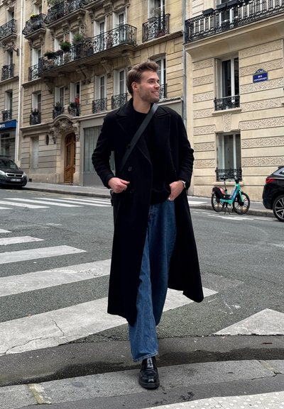 Abrigo largo negro sobre una camisa negra, combinado con jeans anchos azules y zapatos negros, en una calle texturizada con balcones de edificios al fondo.