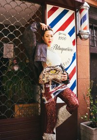 Une femme pose en portant une veste en cuir noire oversize, une robe à motifs avec un design du drapeau britannique, et des leggings en dentelle rouge sophistiquée. Elle tient un sac texturé au motif de serpent.