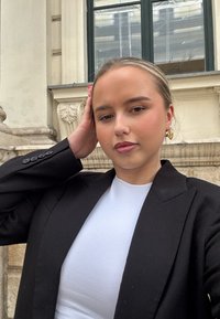 Blazer negro con botones, blusa blanca ajustada, pendientes de aro dorados, piel suave, de pie al aire libre cerca de arquitectura de piedra y ventanas.