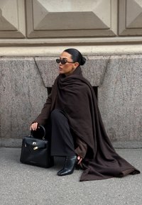 Înveliș maro din lână drapat peste o persoană care poart�ă pantaloni negri și cizme scurte ascuțite. Ține o geantă de piele neagră cu accente aurii.