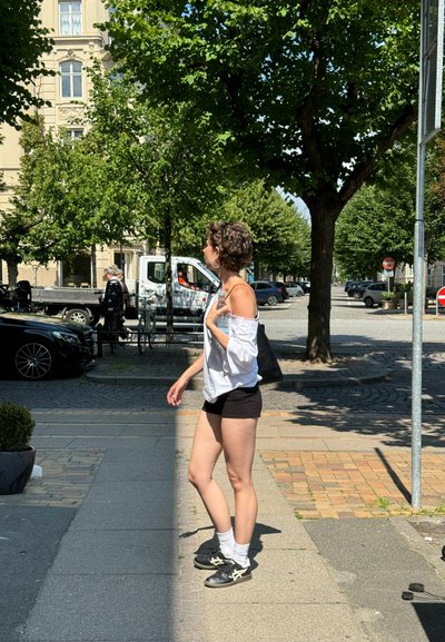 Individuo con una blusa blanca sin hombros y pantalones cortos negros, llevando una bolsa negra. Zapatillas negras con calcetines blancos; escena de calle iluminada por el sol.