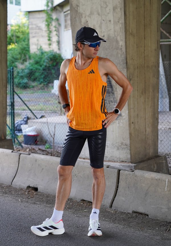 Mees oranži tank-topi, mustade lühikeste pükste, mütsi ja päikeseprillidega seisab silla all, käed puusas, paistab puhkavat pärast trenni.