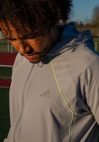 Helehall halli sportjope, millel on õlal geomeetriline muster, helkivad detailid ja triipudel roheline kontrastõmblus.