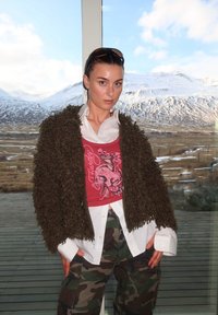 Casaco exterior castanho felpudo sobre uma t-shirt gráfica vermelha, combinado com uma camisa branca de botões e calças camufladas. O fundo apresenta montanhas cobertas de neve.