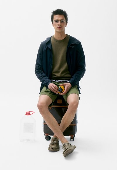 Maleta negra impermeable con acentos marrones, persona vistiendo una chaqueta oscura sobre una camisa verde y pantalones cortos, sosteniendo una cámara amarilla, sentada sobre la maleta.