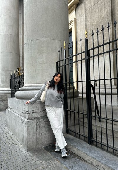 Suéter gris recortado, pantalones blancos con estampado y zapatillas grises. De pie cerca de una columna de piedra y una barandilla negra con acentos dorados.