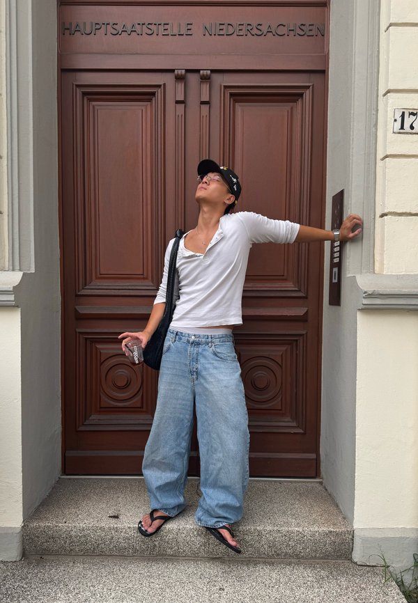 Personne portant une chemise blanche, un jean ample et une casquette noire, appuyée sur un panneau de sonnette devant une grande porte en bois avec le panneau "Niedersachsen".