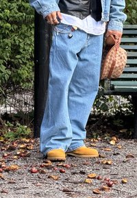Blaue Denim-Jeans mit lockerer Passform, kombiniert mit gelben Stiefeln. Accessoires umfassen eine gemusterte Tasche und Ketten, die vom Hosenbund hängen.
