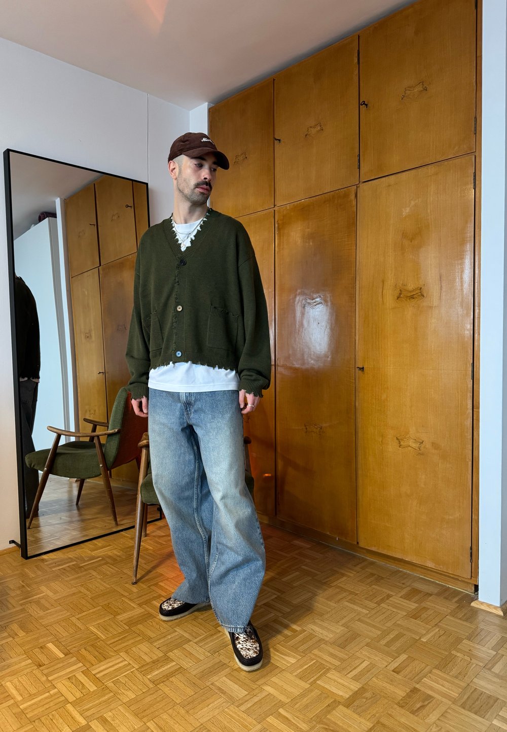 Homme avec moustache portant une casquette marron, un large cardigan vert, une chemise blanche, un jean bleu ample et des chaussures à motifs, debout dans une pièce avec des placards en bois.