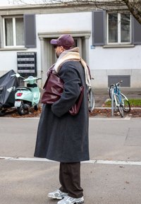 Pitkä harmaa päällystakki, viininpunainen nahkalaukku, beige huivi, tummat housut ja harmaat lenkkarit. Taustalla polkupyöriä ja vihreä skootteri.