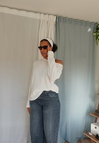 Witte geribbelde off-shoulder trui, relaxed fit, gecombineerd met hooggerokte lichtblauwe jeans. Geaccentueerd met zwarte zonnebril en een witte haarband.