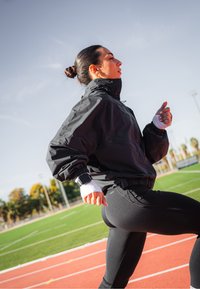 Casaco atlético preto com gola alta, combinado com leggings pretas justas. Detalhes brancos nos punhos e um smartwatch no pulso.