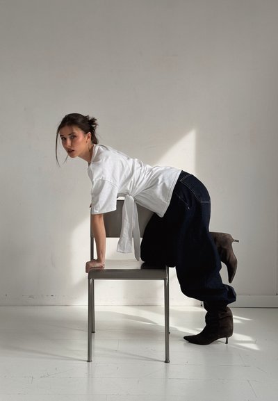 Modelo con una camiseta blanca de gran tamaño y jeans holgados en azul marino, posando sobre una silla de metal gris. Botas de tacón alto marrones completan el conjunto.