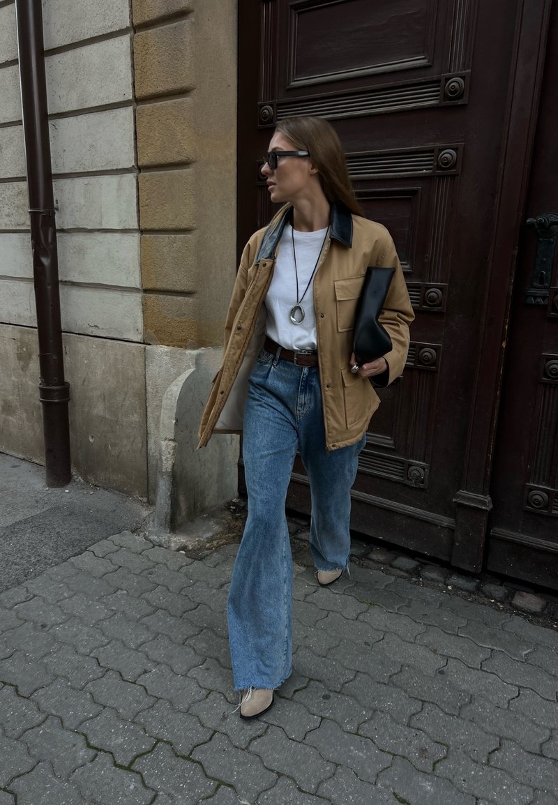 Ανοιχτό καφέ σακάκι με μαύρο γιακά, λευκό μπλουζάκι, ψηλόμεσα μπλε τζιν, μαύρη τσάντα-φάκελος και μπεζ παπούτσια, περπατώντας σε υφαντό πεζοδρόμιο κοντά σε μια πόρτα.