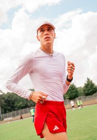 Młody sportowiec w białej koszulce z długim rękawem i czapce, w czerwonych szortach, biegnący po boisku z zamazanymi graczami w tle.