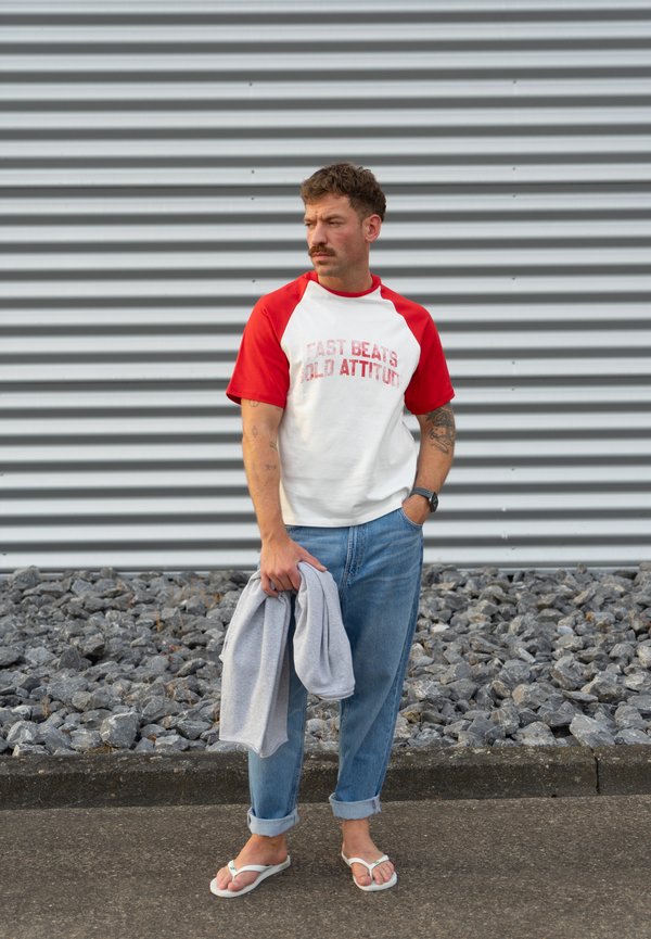 Mann mit Schnurrbart, der ein rot-weißes T-Shirt und blaue Jeans trägt, hält einen grauen Pullover und steht auf einem Gehweg mit Steinen und einer Metallwand im Hintergrund.