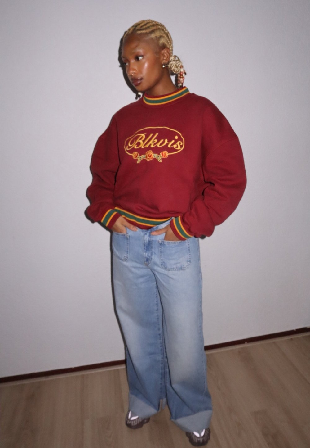 Sweatshirt grená com bordado amarelo e punhos listrados multicoloridos, combinado com jeans de perna larga em denim claro e ténis cinzentos.