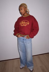 Sweatshirt grená com bordado amarelo e punhos listrados multicoloridos, combinado com jeans de perna larga em denim claro e ténis cinzentos.