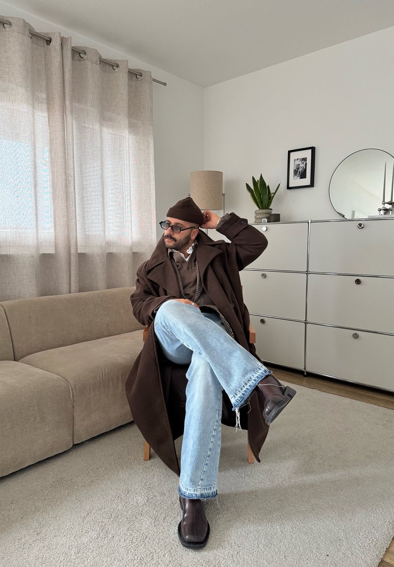 Man with glasses and beard wearing brown coat, beanie, and blue jeans sitting cross-legged on a wooden chair in a neutral-toned living room.