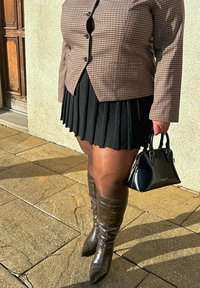 Personne portant un blazer marron à carreaux, une jupe plissée noire, des bottes marron montant jusqu'aux genoux, tenant un sac à main noir Lancaster, debout sur un pavé en pierre.