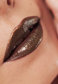 Metaalachtige bronzen lippenstift aangebracht op volle lippen, met een glanzende textuur en reflecterende glans, tegen een achtergrond van een natuurlijke huidskleur.