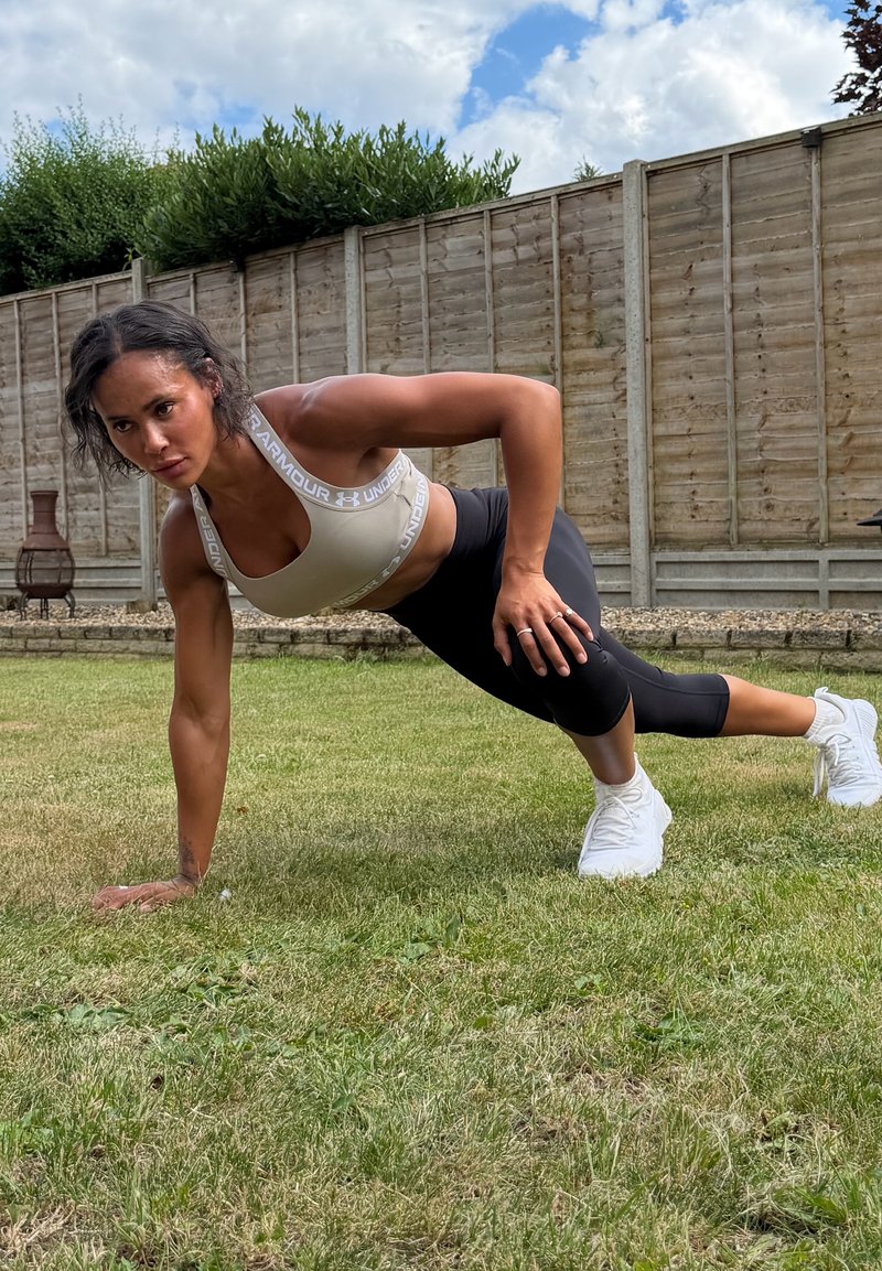 Femme en tenue de sport tenant une position de planche avec un bras et une jambe tendus sur l'herbe dans un jardin clôturé sous un ciel nuageux.
